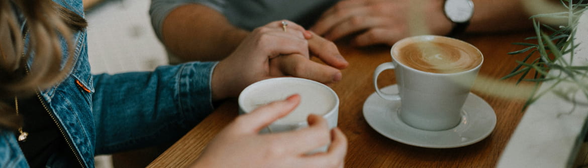 Twee personen in gesprek aan een houten tafel met koffie in een gezellige caféomgeving.