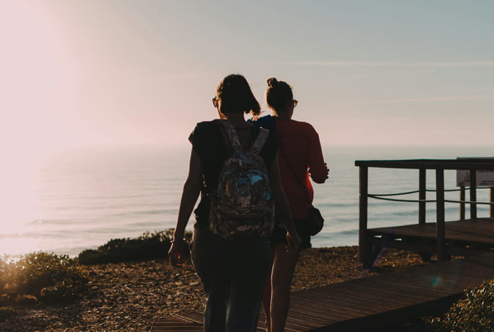 Deux personnes marchent sur un sentier en bois avec vue sur la mer au coucher du soleil.
