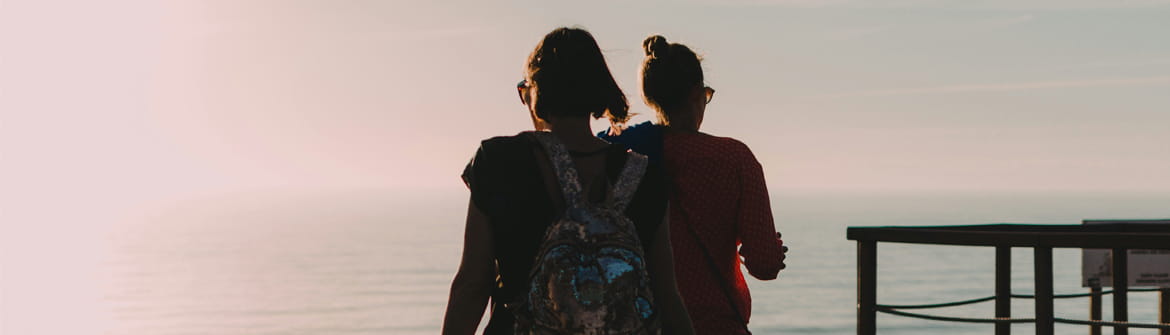 Deux personnes marchent sur un sentier en bois avec vue sur la mer au coucher du soleil.