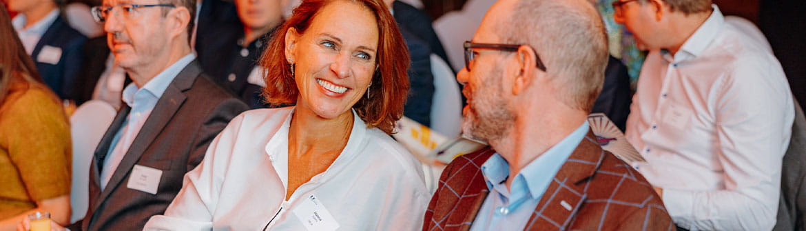 Collega’s zitten naast elkaar tijdens een bijeenkomst en voeren een gesprek in een professionele setting met andere aanwezigen op de achtergrond.