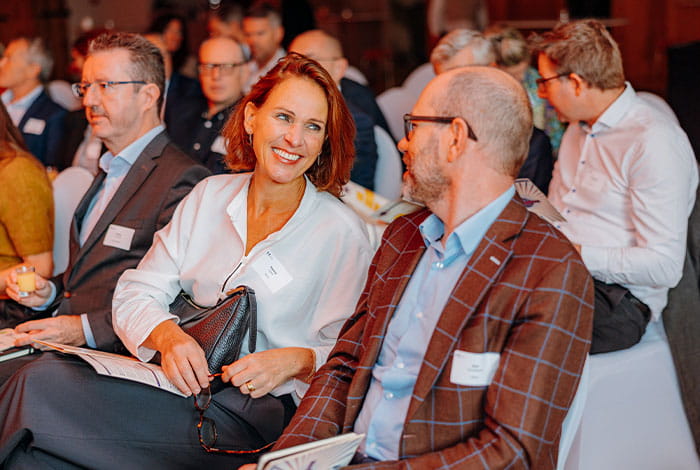 Collega’s zitten naast elkaar tijdens een bijeenkomst en voeren een gesprek in een professionele setting met andere aanwezigen op de achtergrond.
