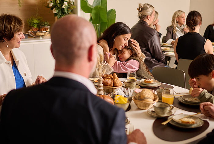 Een groep mensen zit samen aan een gedekte tafel met broodjes, zoetigheden, drank en servies tijdens een gezamenlijke maaltijd.