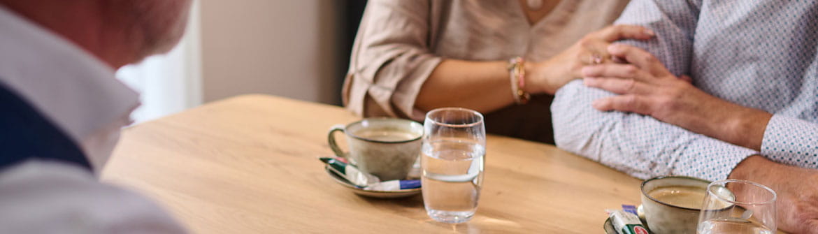Twee personen zitten aan een tafel in gesprek met een uitvaartmedewerker. Op tafel zie je documenten, koffiekopjes en glazen water, terwijl een hand steunend op een arm rust.