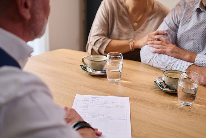 Deux personnes sont assises à une table en entretien avec un collaborateur funéraire. Sur la table se trouvent des documents, des tasses de café et des verres d’eau, tandis qu’une main repose en signe de soutien sur un bras. 