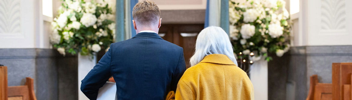  Twee personen lopen samen door een uitvaartzaal met houten banken en grote bloemstukken aan weerszijden.