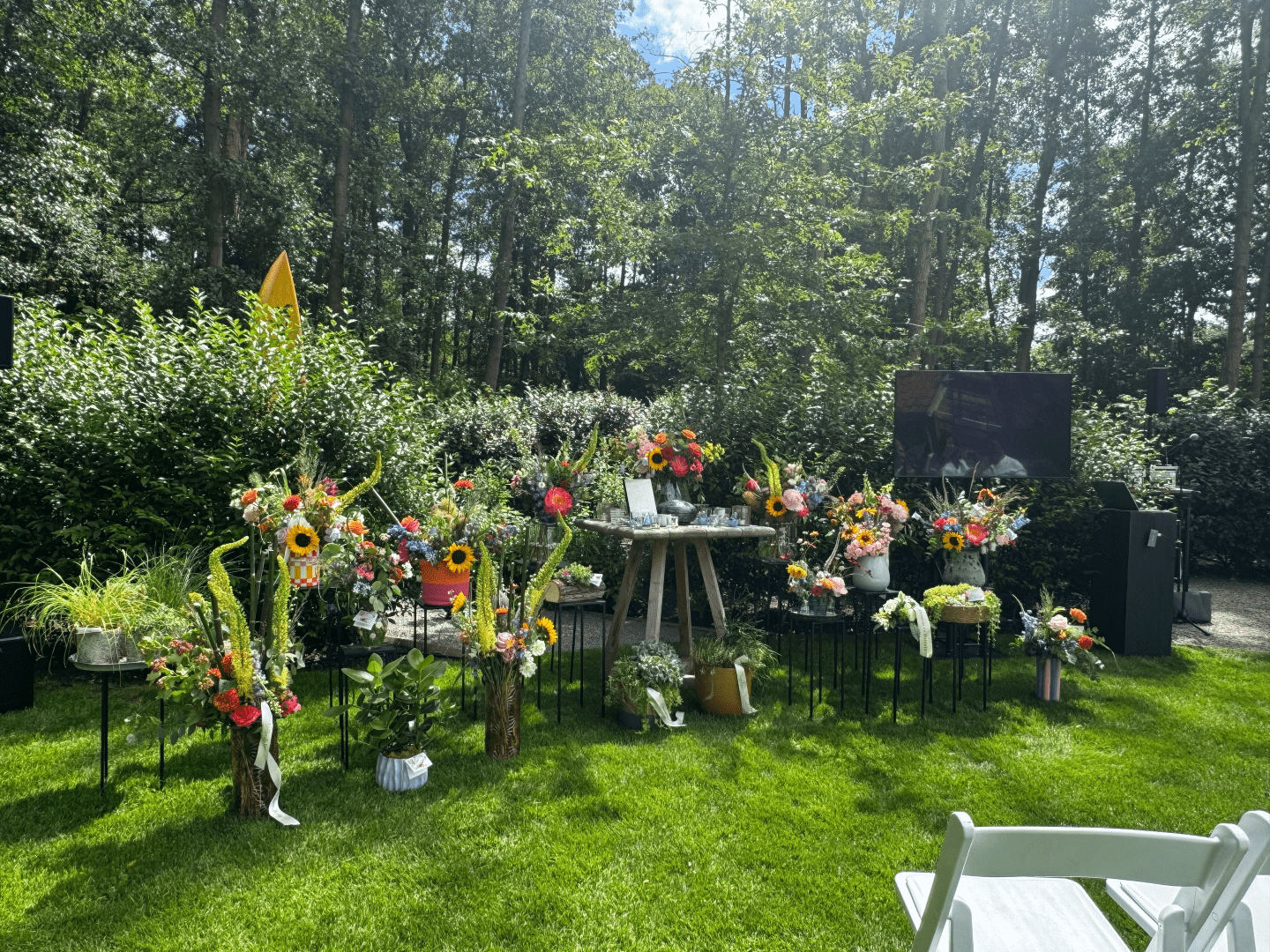 Persoonlijk ingerichte afscheidsplek in een groene tuin. Op het gras staan tientallen kleurrijke bloemstukken rond een centrale herdenkingstafel. Rechts zijn een scherm en geluidsinstallatie opgesteld voor een uitvaartceremonie in openlucht. Op de voorgrond staan witte stoelen voor de aanwezigen. Op de achtergrond vormen bomen en struiken een rustige, natuurlijke omgeving.