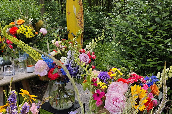Kleurrijke bloemstukken en veldbloemen vormen een persoonlijk herdenkingshoekje in een groene tuinomgeving. Tussen de bloemen staat een gele surfplank als opvallend eerbetoon tijdens een afscheidsplechtigheid in openlucht.