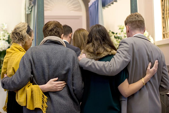 Vijf personen staan dicht bij elkaar in een ruimte, met armen om elkaars schouders, terwijl ze naar voren gericht staan richting een lichte ruimte met bloemen.