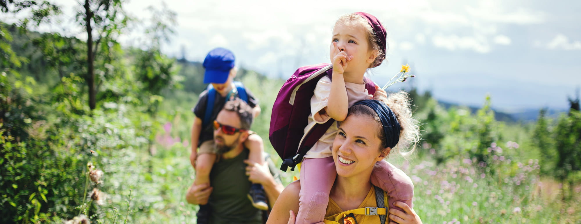 Deux adultes se promènent avec deux enfants sur un sentier naturel ; un enfant est assis sur les épaules avec un sac à dos, tandis que l’autre enfant est également porté.