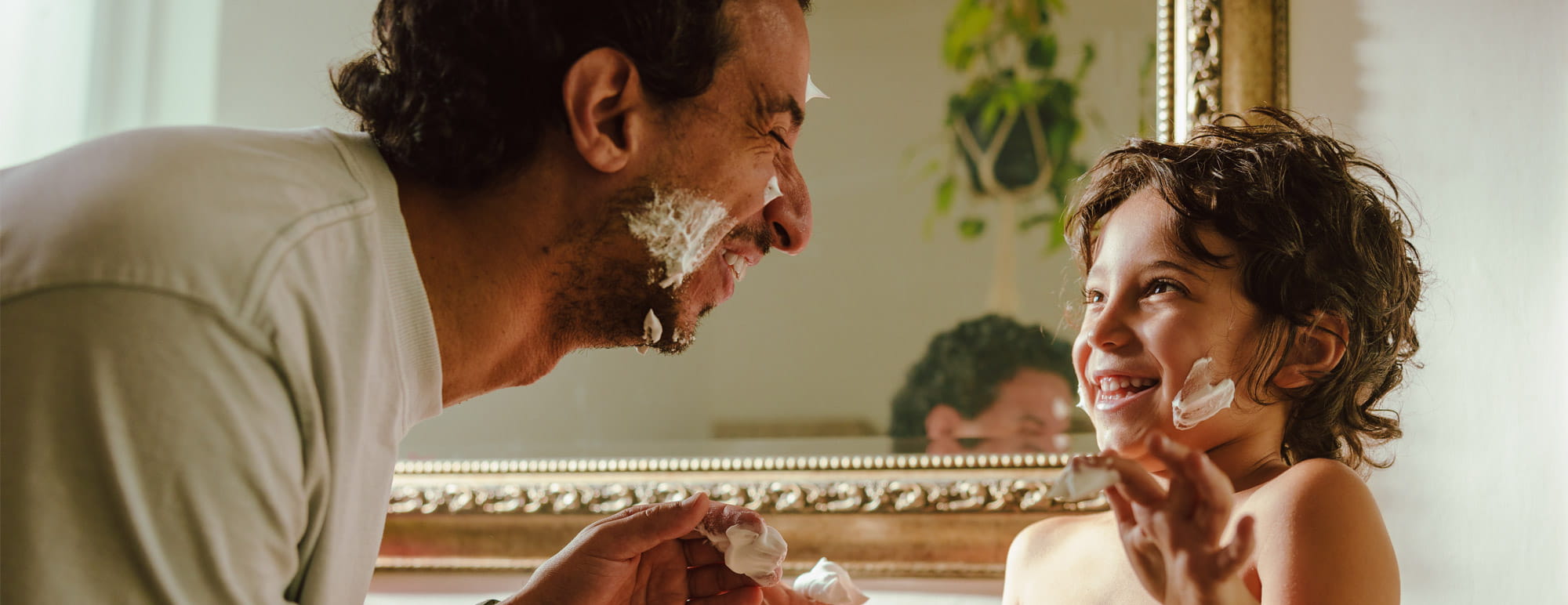 Un adulte et un enfant sont à l’intérieur et s’appliquent de la mousse à raser sur le visage devant un miroir.