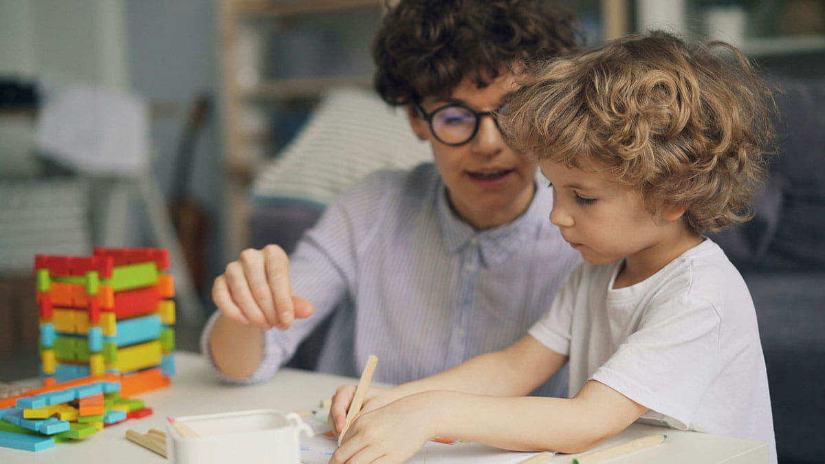Un enfant est assis à une table en train de dessiner tandis qu’un adulte l’accompagne et le soutient.