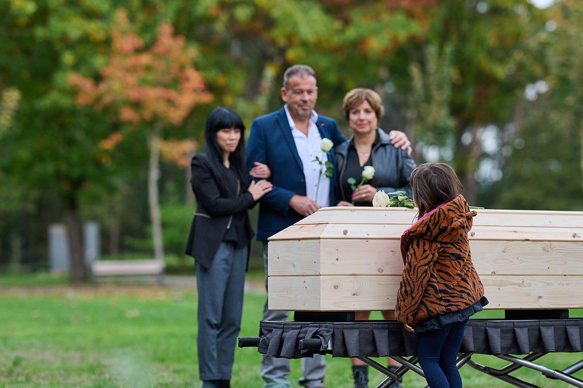 Kind staat bij een houten kist tijdens een afscheidsmoment in een groene buitenomgeving.