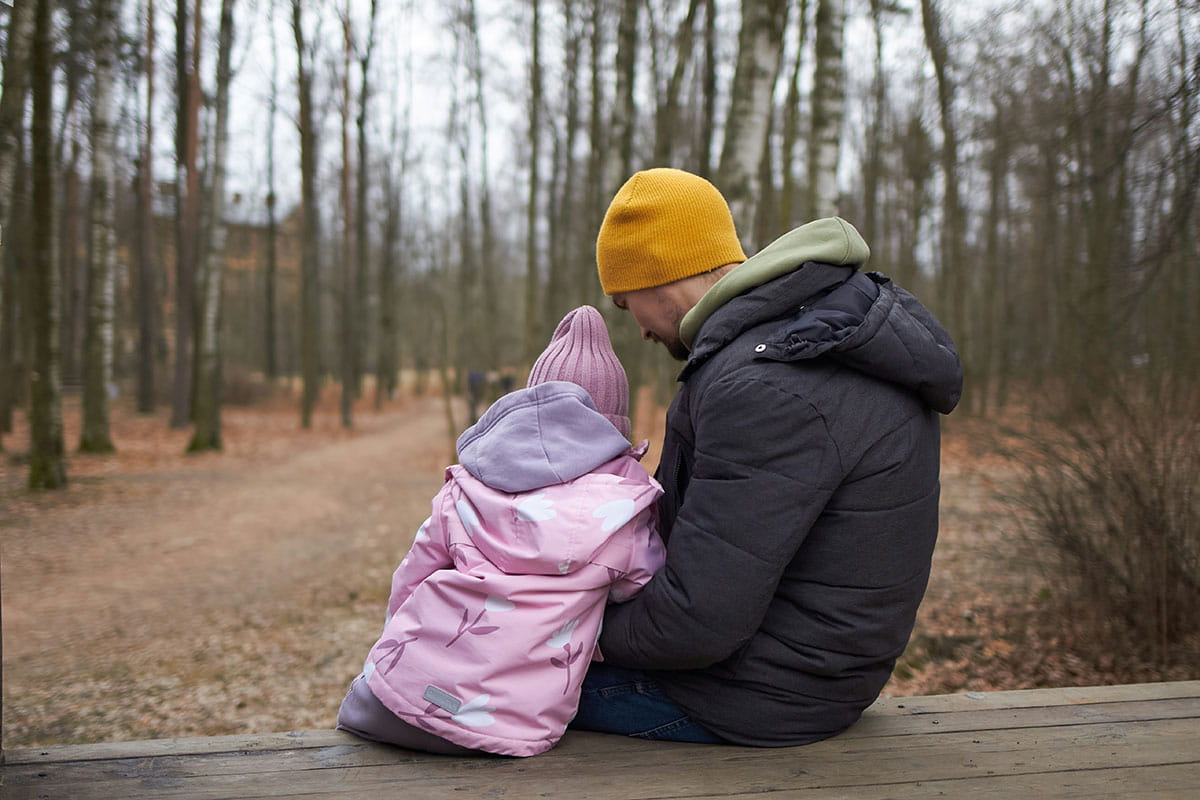 Volwassene en kind zitten naast elkaar op een houten bank in een bosrijk natuurgebied. Ze kijken uit over een wandelpad tussen de bomen terwijl de volwassene een arm om het kind heeft.