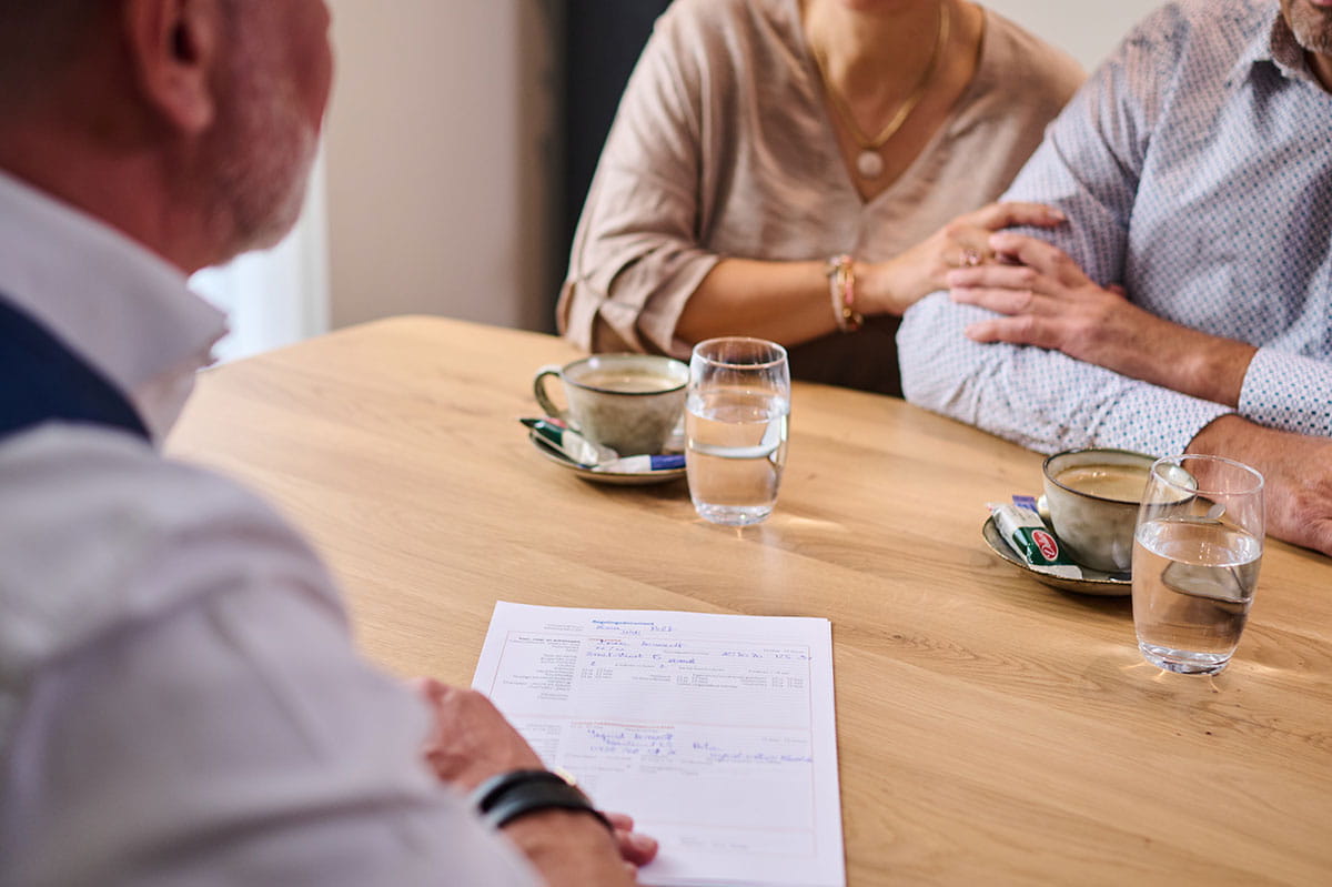 Entretien à table entre un conseiller funéraire et deux personnes, avec des documents, du café et des verres d’eau sur la table.