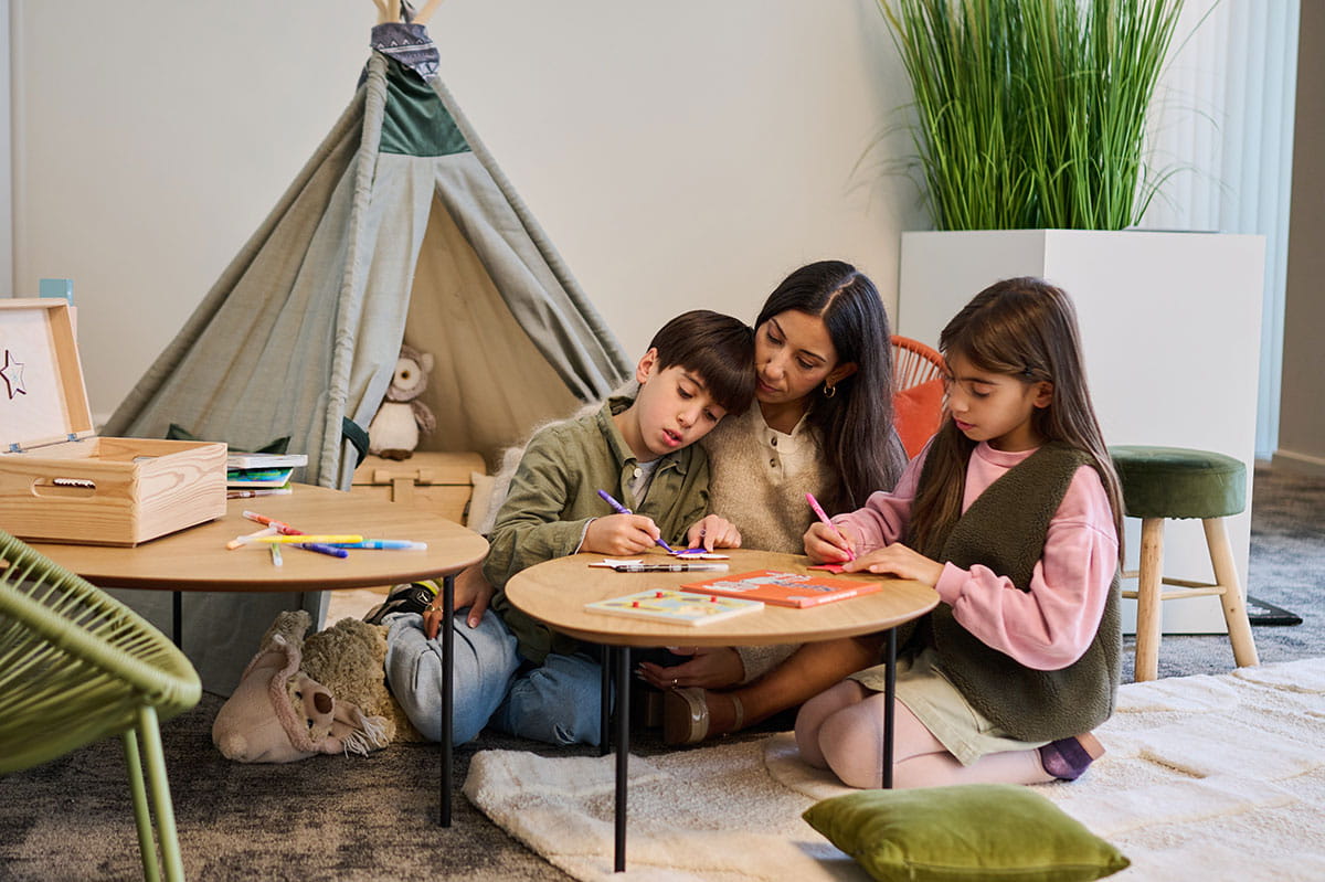 Un adulte et deux enfants colorient ensemble autour de petites tables dans un espace adapté aux enfants.